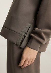 Brązowy sweatshirt o gładkiej teksturze, luźnym kroju i unikalnym detalem w postaci związania na rękawie, umożliwiającym regulację stylu.