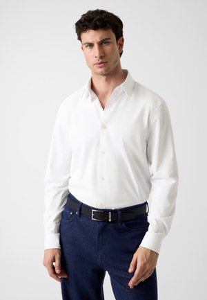 Junger Mann trägt ein weißes Hemd, in dunkelblaue Jeans mit schwarzem Gürtel gesteckt, steht vor einem schlichten hellen Hintergrund.