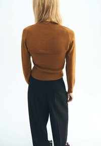 Eine blonde Person steht mit dem Rücken zur Kamera, trägt ein enganliegendes braunes Langarmoberteil und lockere schwarze Hosen vor einem schlichten weißen Hintergrund.
