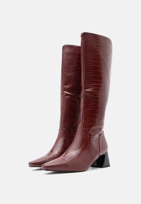 Μποτάκια μπορντό μέχρι το γόνατο με υφή κροκόδειλου, μυτερή μύτη και χοντρό μαύρο τακούνι. Λεπτό φινίρισμα και κομψός σχεδιασμός.