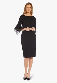 Robe noire fourreau avec un décolleté bateau et des manches cloche transparentes. Coupe ajustée, longueur au genou, accompagnée de talons aiguilles noirs.