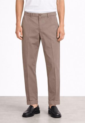 Mężczyzna w beżowych chinosach z mankietami i czarnych skórzanych loafersach, stojący na jasnym tle.