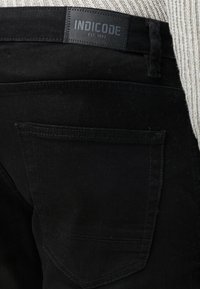 Primo piano della tasca posteriore di jeans in denim nero con etichetta del marchio Indicode cucita in vita su uno sfondo di tessuto chiaro e strutturato.