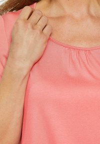 Korte mouwen, top met ronde hals in koraalroze stof met een zachte textuur en geplooide details bij de halslijn, gedeeltelijk bedekt door een hand.