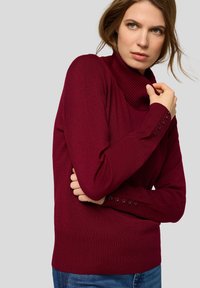 Bordeauxroter Rollkragenpullover mit geripptem Kragen und Bündchen, figurbetontem Design mit Knopfakzenten an den Ärmeln, weichem Stoff und sauberer, gleichmäßiger Textur.