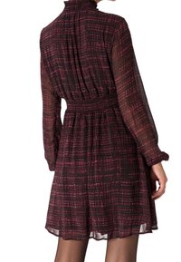 Femme portant une robe longue à manches longues en tissu transparent, avec une taille smockée et un col montant, dans un motif à carreaux rouge foncé et noir.