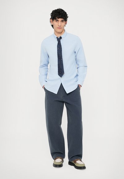 Ανοιχτό μπλε πουκάμισο με κουμπιά συνδυασμένο με σκούρο μπλε φαρδύ παντελόνι. Μαύρη γραβάτα με πολύχρωμες κουκκίδες. Καφέ και λευκά παπούτσια με παχιά σόλα.