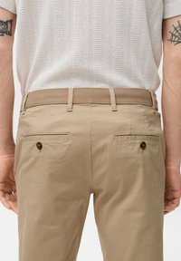 Chino beige con una texture liscia, caratterizzati da un davanti piatto, passanti per cintura e due tasche posteriori con chiusura a bottone. Camicia in maglia bianca visibile sopra.