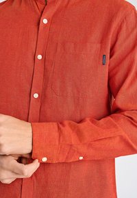 Camicia rossa di lino con colletto button-down, taschino sul petto e polsini regolabili. Presenta una texture sottile e bottoni color crema.