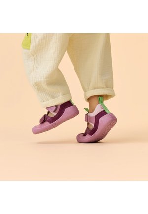 Bambino con pantaloni crema e scarpe rosa-viola con linguette verdi, in piedi sulle punte dei piedi su uno sfondo beige.