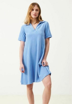 Kvinde iført en lyseblå kjole med korte ærmer og krave, stående foran en ensfarvet hvid baggrund, med hånden på kjolens kant.