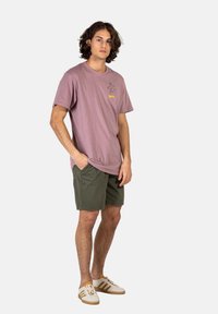 Camiseta de algodón rosa claro con un diseño en la parte frontal, combinada con pantalones cortos verde oliva. El modelo lleva zapatillas blancas con detalles marrones.