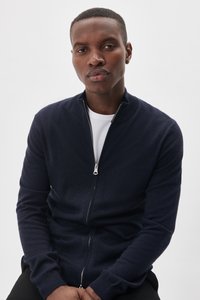 Marinblå zip-up cardigan tillverkad av mjuk stickad tyg, med ståkrage och långa ärmar. Den har en silverfärgad dragkedja och ribbade ärmmuddar.