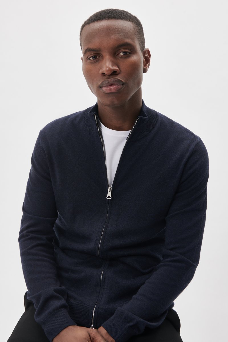Marinblå zip-up cardigan tillverkad av mjuk stickad tyg, med ståkrage och långa ärmar. Den har en silverfärgad dragkedja och ribbade ärmmuddar.