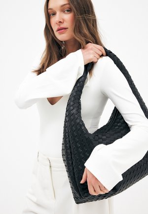 Pikkade juustega naine, kes kannab valget topi ja pükse, hoiab õlal suurt musta punutud käekotti ühevärvilise tausta ees.