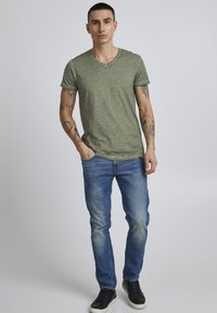 Zeleni heathered V-izrez majica napravljena od mekog materijala, u kombinaciji s klasičnim plavim trapericama. Model nosi crne tenisice, a tetovaže su vidljive na rukama.