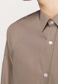Brown, button-up shirt featuring a smooth texture, pointed collar, and light buttons. Close-up view of collar and part of the sleeve.
