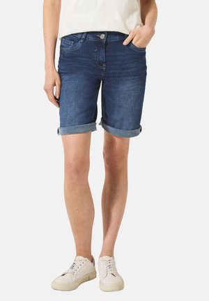 Shorts en denim bleu foncé avec des poignets retroussés, dotés d'une taille avec un bouton, de cinq poches et d'une coupe classique. Portés avec des baskets blanches.