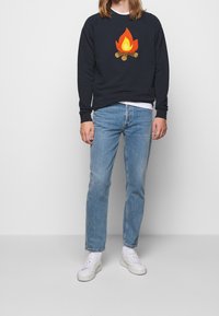 Mörkblå sweatshirt med en grafik av en lägereld, kombinerad med ljusblå raka jeans och vita sneakers. Tyget ser mjukt ut med en avslappnad passform.