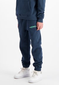 Marineblauwe sweatpants met elastische boorden, voorzien van een groen grafisch logo op de linkerdij. Het materiaal lijkt zacht en heeft een relaxte pasvorm.