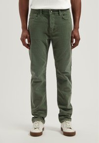 Pantalones de pana verde de corte ajustado, con cinco bolsillos y cremallera. Llevados con zapatillas blancas, mostrando una tela texturizada.