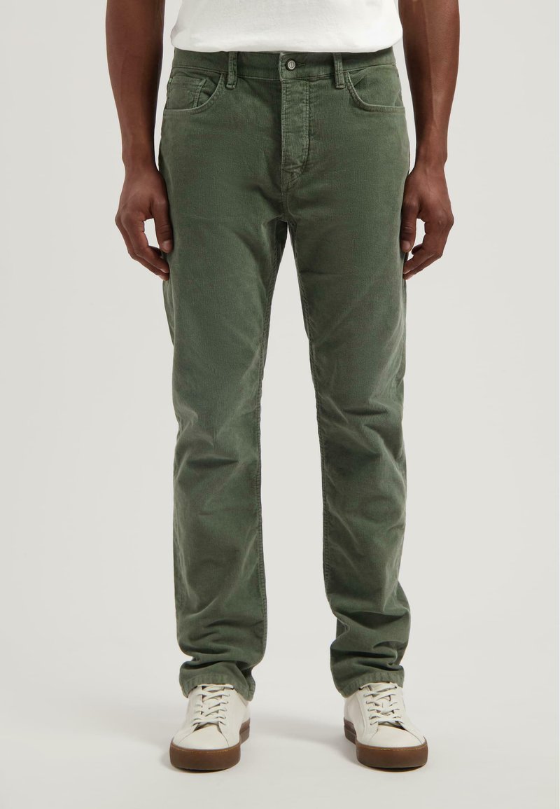 Pantalones de pana verde de corte ajustado, con cinco bolsillos y cremallera. Llevados con zapatillas blancas, mostrando una tela texturizada.