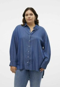 Camisa de mezclilla en un tono azul medio, con un cierre frontal de botones, mangas largas y un corte relajado con un dobladillo curvado. Textura suave.