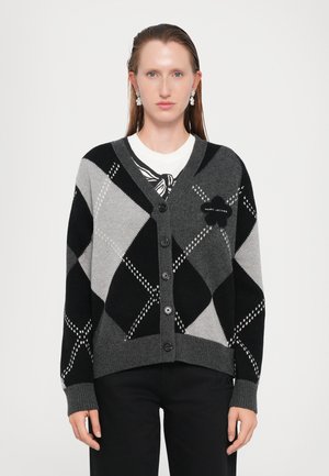 Kvinne i svart og grått rutet cardigan med knapper, blomsterapplikasjon og hengende øredobber, står foran en ensfarget lys bakgrunn.