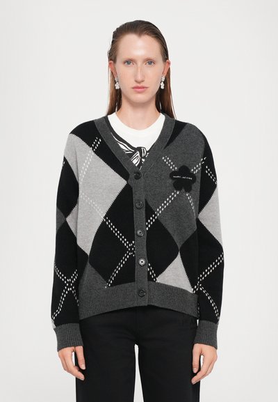Donna che indossa un cardigan a bottone argyle nero e grigio con una toppa a forma di fiore e orecchini a goccia, in piedi di fronte a uno sfondo chiaro e uniforme.