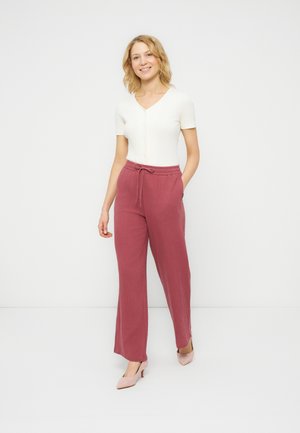 Femme souriante debout, les mains dans les poches, portant un haut blanc à manches courtes, un pantalon ample mauve avec cordon de serrage, et des chaussures à talons roses, sur fond blanc.