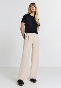 Giovane donna con lunghi capelli biondi che indossa una maglietta nera ricamata, pantaloni beige a gamba larga e zoccoli neri, in piedi di fronte a uno sfondo neutro.