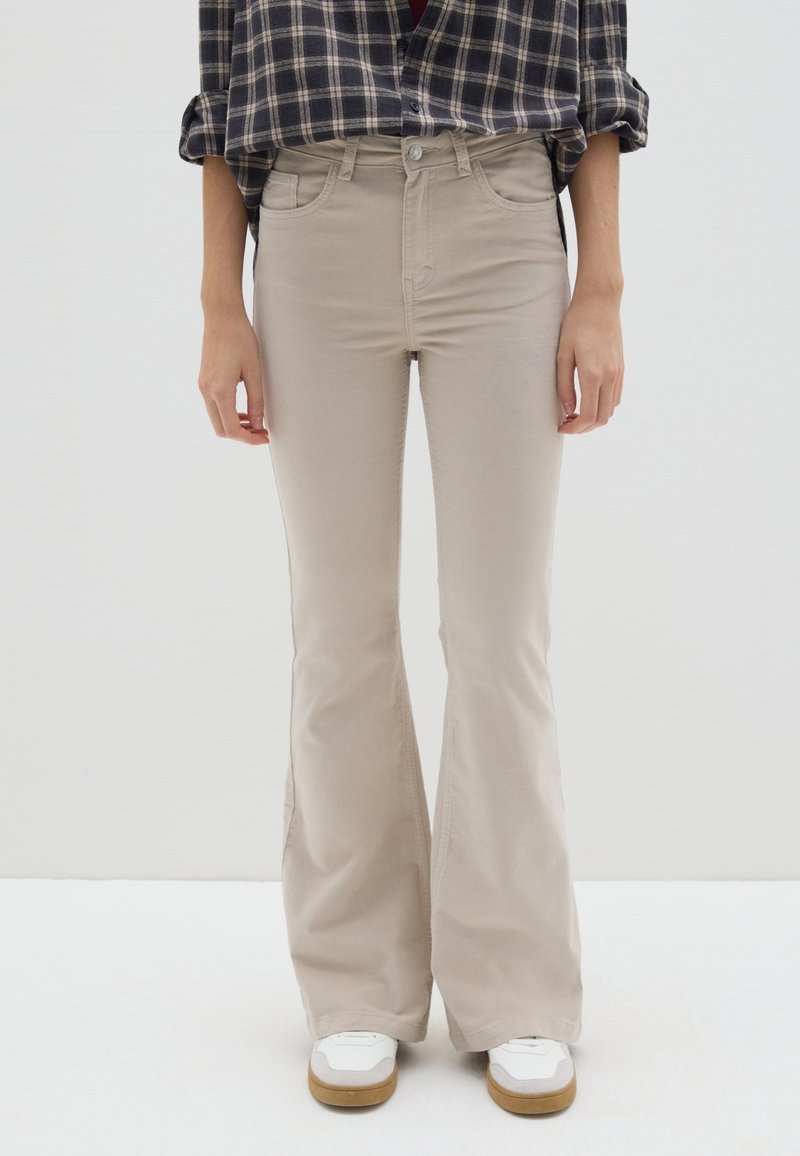 Beige flare-broek met een gladde textuur, hoge taille en zijzakken, gekoppeld aan witte sneakers met een lichtbruine zool.