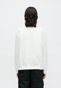Pessoa com cabelo castanho escuro na altura dos ombros, vestindo um sweatshirt branco simples e calças pretas, de costas contra um fundo branco.