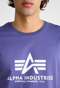 Violet kokvilnas t-krekls ar baltu logo, kas satur leņķīgu dizainu un tekstu "ALPHA INDUSTRIES, KNOXVILLE, TENNESSEE, U.S.A."