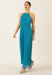 Teal color pleated jumpsuit dengan leher halter dan ikat pinggang elastis, dilengkapi dengan detail rantai di leher. Dipadukan dengan sandal hak tinggi metalik.