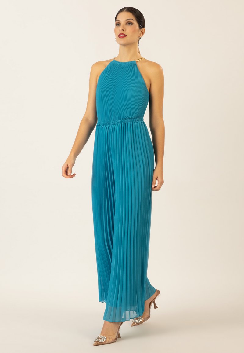 Combinaison plissée teal avec un col halter et une taille élastique, présentant un détail en chaîne au niveau du cou. Associée à des sandales à talons métalliques.