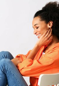 Una donna dai capelli ricci indossa una camicia arancione con bottoni e jeans in denim, seduta con la mano sulla guancia, sorridendo. Sfondo bianco.