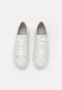 White leather sneakers with a smooth texture, round toe, and flat laces. Features a subtle logo in gold on the tongue and a minimal design.