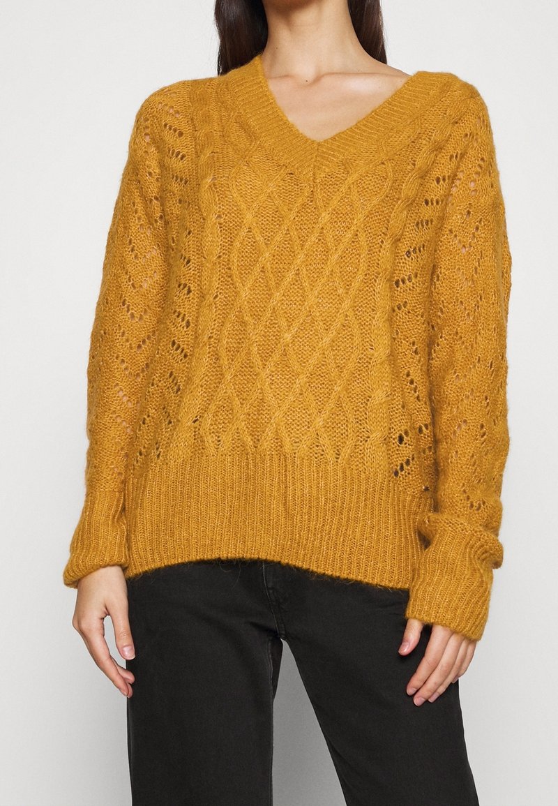 Mosterdgele gebreide trui met een v-hals, voorzien van gestructureerde diamantpatronen en opengewerkte accenten. Ribgebreide manchetten en zoom.