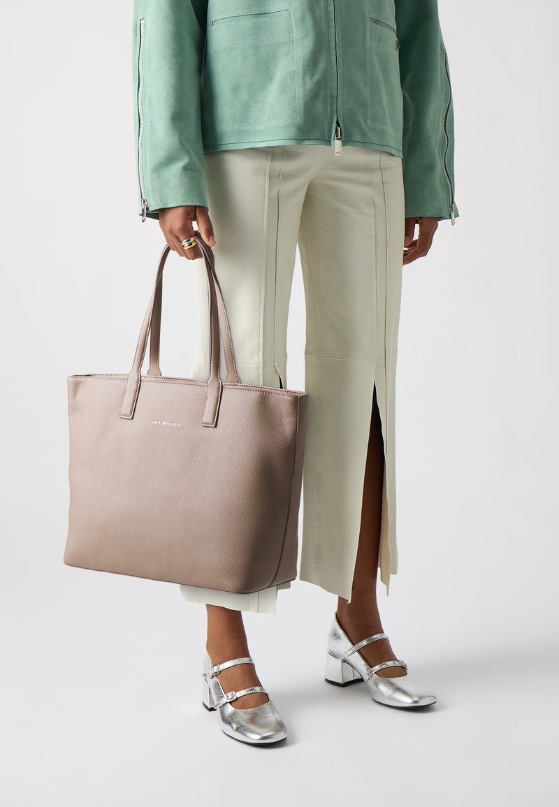 Sac fourre-tout en cuir beige avec deux poignées supérieures, présentant une texture lisse et un design minimaliste. Porté avec une veste vert clair et un pantalon crème.