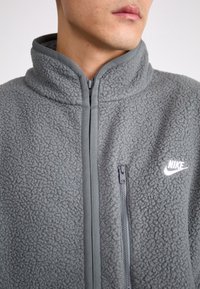 Grå fleecejacka med hög krage, dragkedjeförsedd framficka och Nike-logotyp. Har mjuk textur och svag glans. Dragkedja i kontrasterande grå nyans.