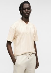 Μπεζ κοντομάνικο πουκάμισο με κάθετες ρίγες, ντεκολτέ σε σχήμα V και κεντρικό έμβλημα. Λείο ύφασμα, συνδυασμένο με ανοιχτόχρωμο παντελόνι με κορδόνι στη μέση.