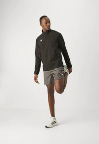 Homme en veste noire, short gris et baskets blanches étirant la jambe droite en tenant le pied derrière lui sur un fond uni.