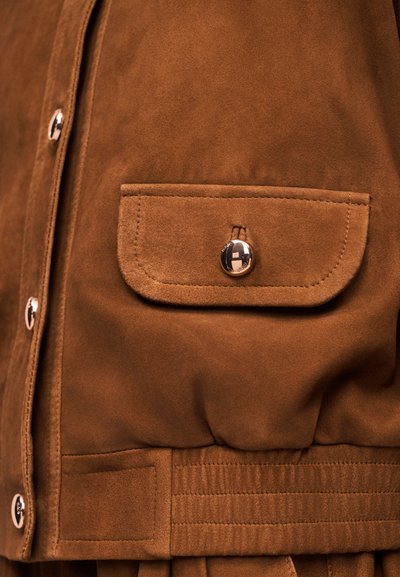 Καφέ σαμουά σακάκι με κουμπωτή τσέπη με καπάκι και ασημένια κουμπιά που τρέχουν κάθετα κατά μήκος του εμπρόσθιου κλεισίματος.