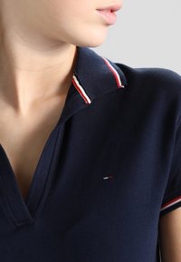 Námorně modré polo tričko s klasickým límcem, zdobené červenými a bílými pruhy. Měkká látka, viditelné švy a malé logo na hrudi.