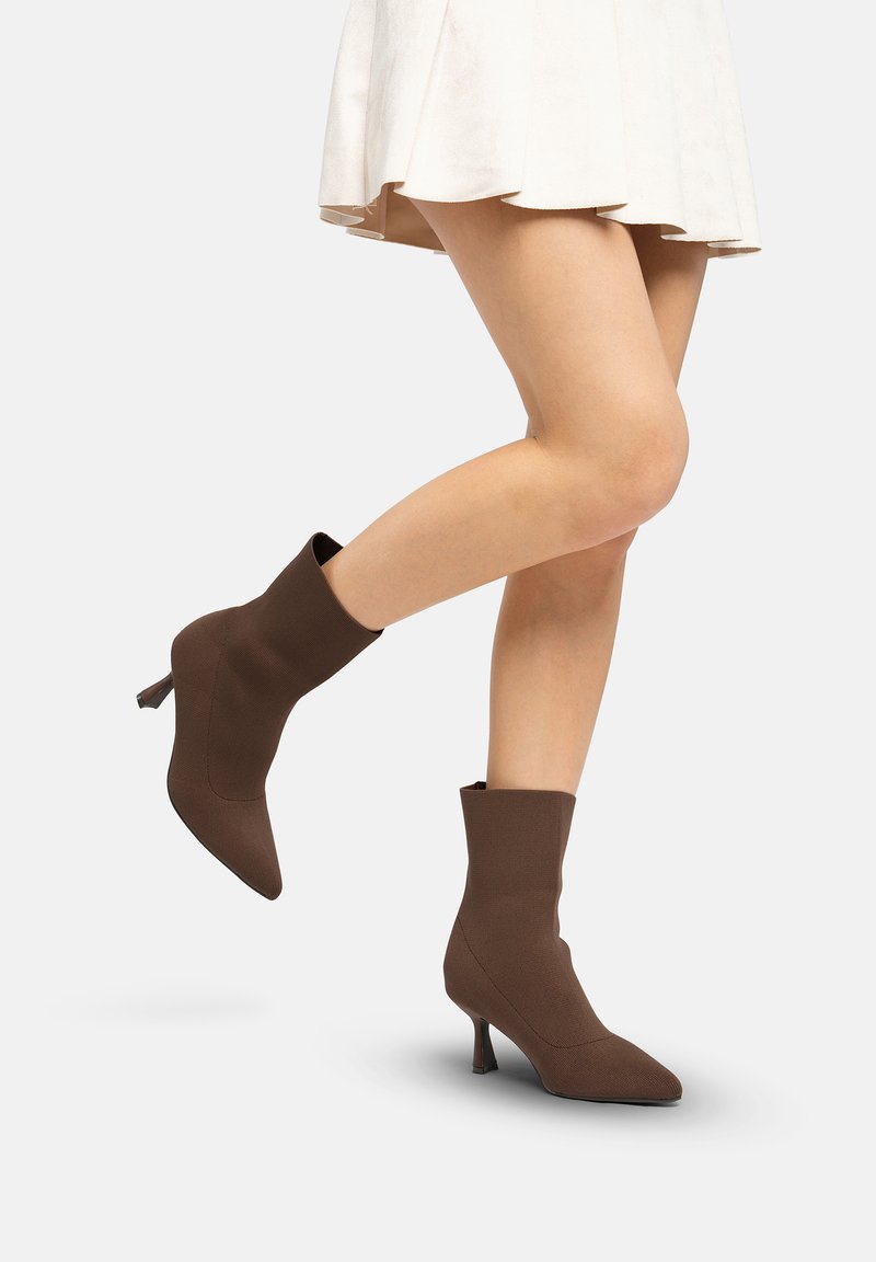 Botas de tobillo marrones tejidas con punta puntiaguda y tacón de cono de 5 cm, con una textura suave y forma ajustada, combinadas con una falda blanca campana.