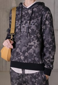 Felpa con cappuccio in un motivo camouflage nero e grigio, dotata di cappuccio regolabile e polsini a costine. Abbinata a una borsa a tracolla beige.