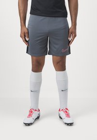 Grå sportshorts med en rosa Nike-logotyp, parat med vita knästrumpor och vita fotbollsskor med röda snörning och svarta detaljer.