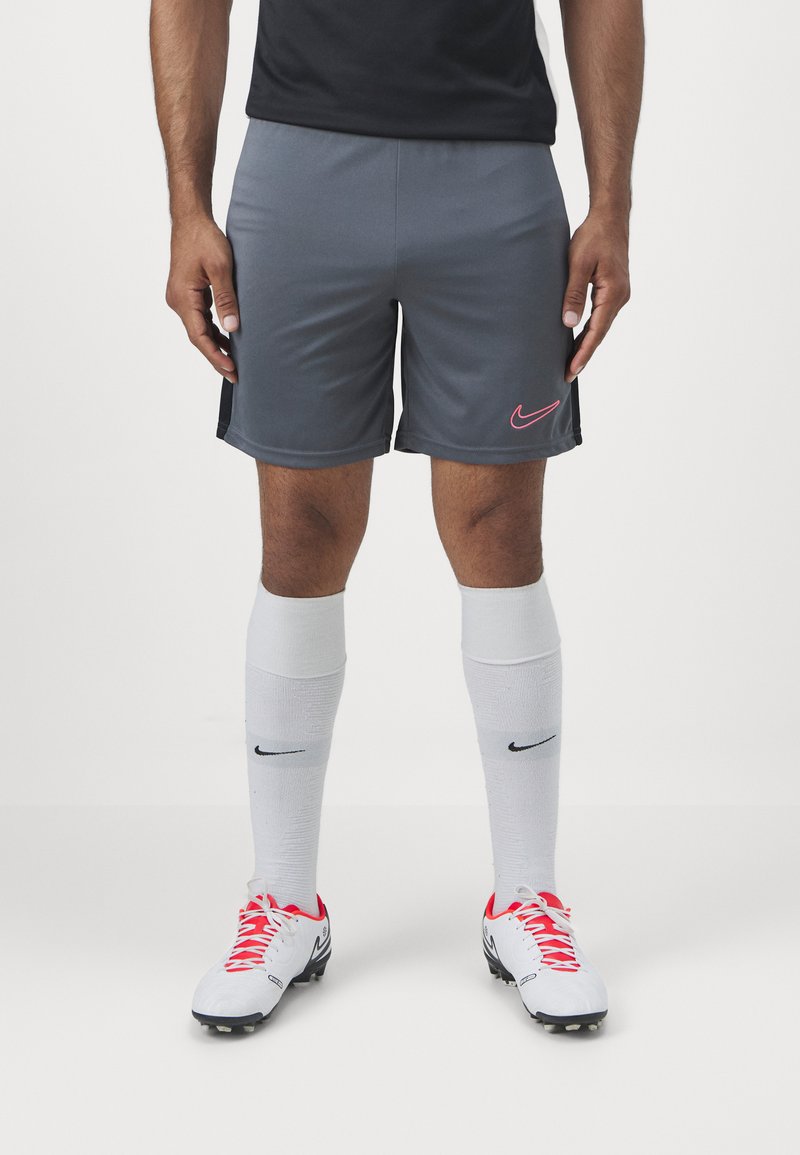 Grå sportshorts med en rosa Nike-logotyp, parat med vita knästrumpor och vita fotbollsskor med röda snörning och svarta detaljer.