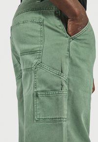 Persoană îmbrăcată cu pantaloni utility verzi, cu mai multe buzunare, având mâna odihnită în interiorul unui buzunar frontal pe un fundal neutru.